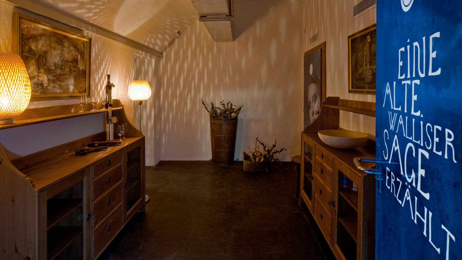 Salle de réception du Johanneli Fi Weinkeller avec des buffets en bois et la lumière des lampes, à droite la porte bleue portant l'inscription «Eine alte Walliser Sage erzählt»