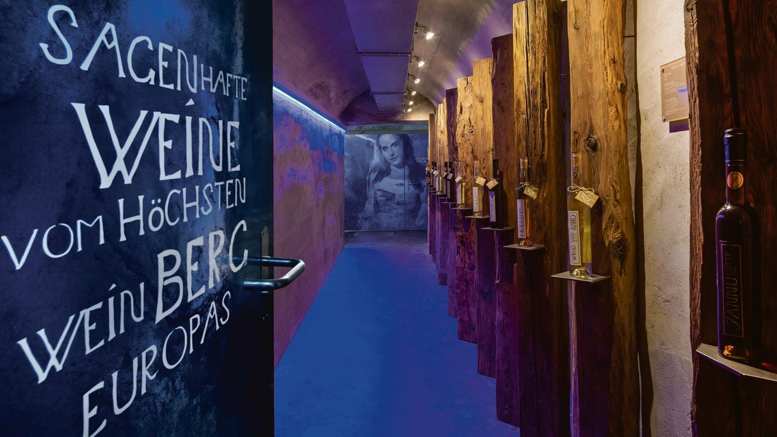 Galerie rocheuse illuminée à l'entrée du Johanneli Fi Weinkeller, à droite, des bouteilles de vin sur de vieilles poutres en bois